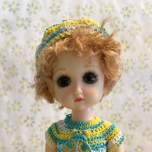 May include: A doll wearing a handmade crochet outfit. The outfit includes a striped yellow, white, and teal top with a ladybug applique, matching hat, and floral print pants. The doll is also wearing crochet shoes.