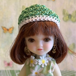 May include: A doll with brown hair wearing a green crocheted hat and a green and white daisy print dress with a yellow crocheted belt.