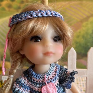 May include: A doll with blonde hair wearing a blue and pink crocheted dress with pink flower accents and a blue and pink braided headband.