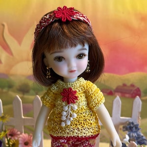 May include: A small doll with brown hair wearing a yellow crocheted top with a red flower design, red and white striped shorts, and red crocheted shoes.