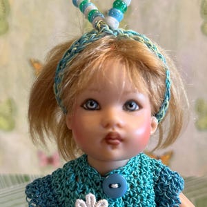 May include: A doll with blonde hair, blue eyes, and rosy cheeks. The doll wears a turquoise crocheted top with a white flower and blue buttons. A matching headband and beaded antennae complete the look.