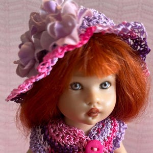 May include: A doll with red hair and blue eyes wearing a crocheted pink, purple, and white outfit. The doll has a matching hat with a pink brim and lavender flower accents. The outfit includes a button closure.