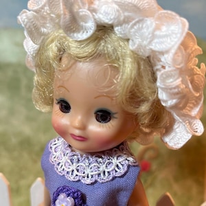 May include: A vintage doll with blonde hair and a white lace bonnet. The doll wears a lavender dress with a white lace collar and a purple flower applique. The doll has rosy cheeks and is set against a blurred background.