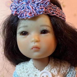 May include: A doll with dark hair and a crocheted headband with a blue and pink flower. The doll is wearing a white crocheted top with blue accents and a large white button.