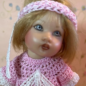 May include: A doll with blonde hair and blue eyes wearing a pink crocheted dress and headband. The doll's dress has white trim and a decorative white section at the neckline. The headband is a braided design.