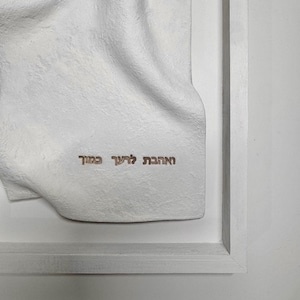 May include: A white, textured wall art piece with a draped fabric design, framed in a white wooden frame. The artwork features Hebrew text in a gold color. The overall aesthetic is minimalist and modern.