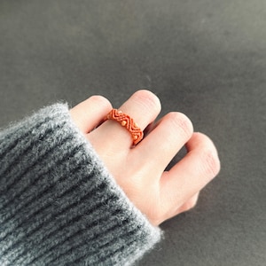 Puede incluir: Primer plano de un anillo de macramé naranja con un patrón en zigzag y pequeños detalles de cuentas. El anillo se lleva en un dedo, con una manga de suéter gris en el fondo. Joyería hecha a mano.
