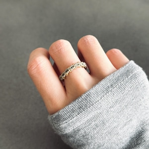 Puede incluir: Un anillo dorado con un patrón repetido de pequeñas piedras brillantes. El anillo se lleva en un dedo, con una camiseta de manga larga gris en el fondo. El diseño del anillo es intrincado y llamativo.