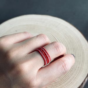 May include: A red ring with a woven design, featuring a band of small, silver-coloured beads. The ring is worn on a finger, set against a wooden background.