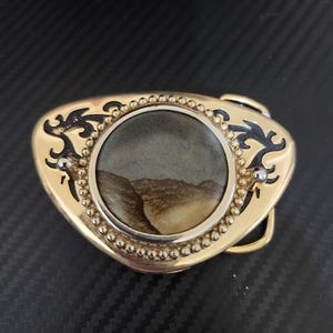 May include: A gold-toned belt buckle with a central circular image of a desert landscape. The buckle has a decorative border with small silver beads and black filigree accents. The buckle is a western-style accessory.