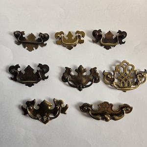 May include: Seven antique brass drawer pulls in different designs are displayed. The pulls have ornate details, such as scrolled edges and floral patterns. The finishes vary from dark bronze to bright gold, providing a vintage aesthetic.