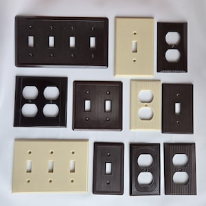 May include: A collection of vintage electrical switch plates in various colors and designs. The plates include toggle switch covers, outlet covers, and a combination of both. Colors range from dark brown to cream and black.