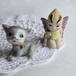 May include: Two porcelain figurines, a grey cat with a red collar and a gold bell, and a yellow fairy with pink wings and a flower.
