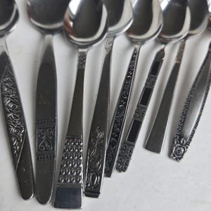 May include: A collection of stainless steel spoons with various handle designs. The spoons have different patterns, including floral, geometric, and textured details. The spoons are arranged in a fan shape, showcasing their diverse styles.