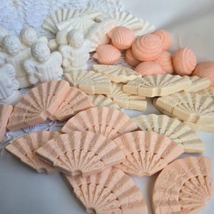 May include: A collection of decorative soaps in different shapes and colours. The image shows white angel figurines, peach-coloured seashell and fan-shaped soaps, and off-white fan-shaped soaps. The soaps are arranged on a white lace surface.