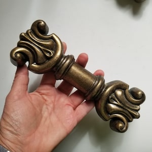 May include: A gold-toned decorative cabinet hardware pull with ornate scrollwork details. The pull is shaped like a stylised letter 'T' with a rounded top and bottom.