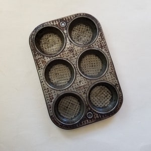 May include: A vintage, well-used six-cup muffin tin with a distressed, speckled finish. The metal pan has a rectangular shape with rounded cups and the words "BAKE-RITE" and "NY, NY" are visible. The pan has a small logo and the letter "G".