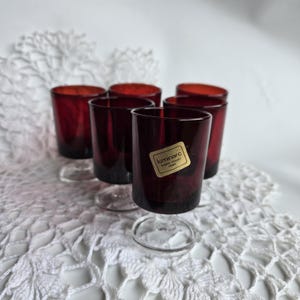 May include: Six ruby red Luminarc glasses with clear stems are arranged on a white lace doily. The glasses have a short, cylindrical shape and a dark red hue. A gold sticker is visible on one glass.