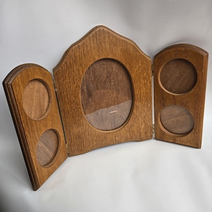 May include: A wooden, tri-fold picture frame with an oval centre and two sets of two circular openings on the side panels. The frame is made of light brown wood and has a natural wood grain.