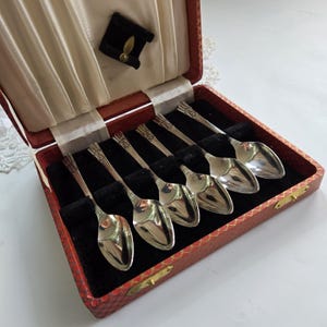 May include: A set of six silver-toned metal spoons with ridged handles, arranged diagonally in a rectangular red textured case. The interior of the case features a black velvet-lined base with individual slots for each spoon and a cream-coloured fabric lid.