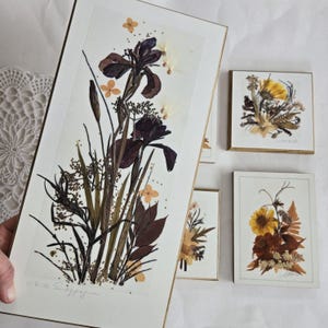 May include: Four framed pressed flower art pieces. The largest piece features a purple iris with brown stems and leaves. The smaller pieces feature yellow and brown flowers with green stems and leaves. The largest piece is signed "ela.m. swayze".
