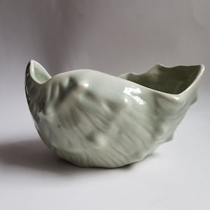 May include: A pale green ceramic bowl with a textured, organic design. The bowl has a unique, asymmetrical shape with a flared rim and a glossy finish. The bowl's surface has a rippled texture, adding visual interest.