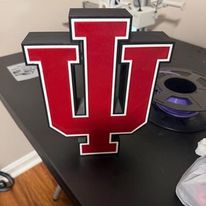 Peut inclure: Un logo tridimensionnel de l'Université de l'Indiana présentant les lettres I et U rouges entrelacées, soulignées de blanc et encadrées de noir. L'emblème est construit avec une conception en couches, affichant une finition rouge foncé.