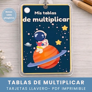 Puede incluir: Un llavero de tablas de multiplicar con temática espacial. El diseño presenta un astronauta leyendo un libro mientras está sentado en un planeta, con estrellas y un OVNI en el fondo. Se muestra el texto "Mis tablas de multiplicar".