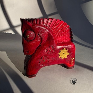 May include: A carved wooden horse figurine in a vibrant red hue, adorned with gold star and sunburst accents. The horse features detailed carving and brass-colored studs, showcasing a folk art style.