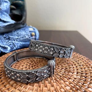 May include: Two silver-toned bangles with intricate leaf and floral patterns. The bracelets are displayed on a woven, light brown surface. The bangles have a vintage or antique aesthetic, with detailed metalwork and a slightly darkened finish.