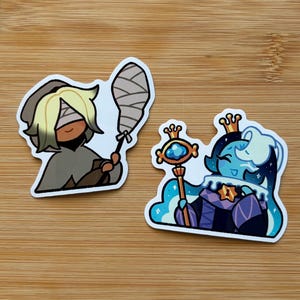 May include: Two stickers featuring cartoon characters. One is a figure with blonde hair, bandages, and a gray cloak, holding a wrapped item. The other is a blue character with a crown and scepter, wearing a purple and blue outfit.