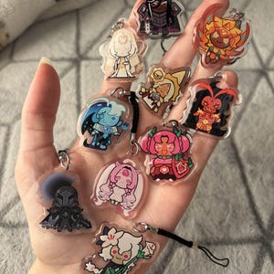 May include: A collection of colorful acrylic charms featuring various cartoon characters. Each charm has a unique design with vibrant colors and attached keychains. The charms are held in a hand, showcasing their size and detail.