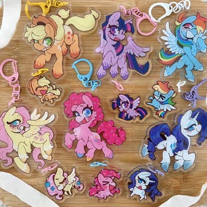 May include: A collection of clear acrylic keychains featuring colourful cartoon pony characters with various expressions and poses. Each charm includes a plastic lobster clasp in matching colours like yellow, pink, blue, white, and purple, showcasing detailed character illustrations.