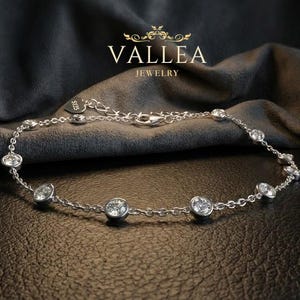 May include: A silver bracelet with clear, round gemstones evenly spaced along a delicate chain. The bracelet is displayed on a dark, textured surface with a soft, dark gray fabric in the background. The brand name "VALLEA JEWELRY" is visible.