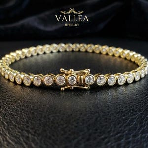 Op de afbeelding: Een gouden armband met ronde, heldere edelstenen, gezet in individuele zettingen. De armband heeft een sluiting en wordt gepresenteerd op een zwarte ondergrond. De merknaam "VALLEA JEWELRY" is zichtbaar.