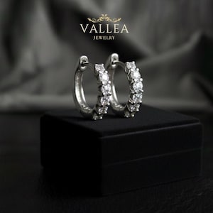 Può includere: Un paio di orecchini a cerchio in argento con diverse pietre preziose trasparenti. Gli orecchini sono esposti su una scatola nera contro uno sfondo scuro e sfocato. Il marchio "VALLEA JEWELRY" è visibile.