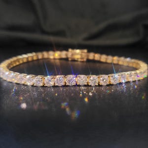 May include: A gold tennis bracelet with numerous round-cut, clear gemstones. The bracelet is set on a dark surface, with the Vallea Jewelry logo in the upper left corner. The gemstones sparkle, reflecting light.