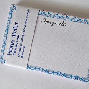 Puede incluir: Un bloc de notas blanco con la palabra "Margarita" en escritura cursiva. El bloc de notas está envuelto con una banda que dice "Prana Atelier Thank you cards" y tiene un borde azul y rojo.