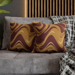 May include: Two square throw pillows with a swirling pattern in shades of burgundy, gold, and cream. The pillows are placed on a gray sofa, with a patterned blanket visible in the background. The pillows add a decorative touch to the living room.