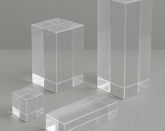 May include: Four clear acrylic display blocks of varying sizes. The blocks are rectangular and square, with a transparent appearance. The arrangement includes a small cube, a long rectangle, and two taller rectangular blocks, all on a gray surface.