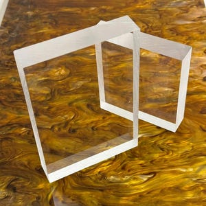 May include: Two transparent, rectangular acrylic blocks are placed on a surface with a swirling pattern of gold, brown, and yellow hues. The blocks are clear, allowing the background to be partially visible.