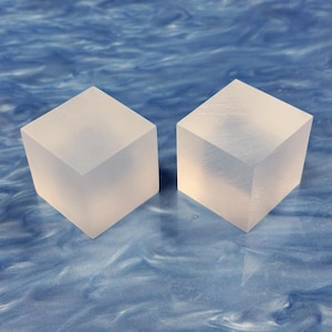 May include: Two translucent, square cubes sit side-by-side on a blue surface. The cubes are made of a frosted material, and each has six visible sides. The cubes are evenly lit, and the edges are sharp and defined.