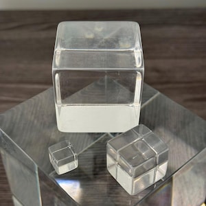 Puede incluir: Tres cubos de acrílico transparente de diferentes tamaños. El cubo más grande está sobre una superficie clara e inclinada. También se ven un cubo más pequeño y un cubo estilo Rubik. Los cubos son transparentes.