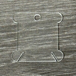 May include: A clear acrylic spool-shaped craft supply with a square center and rounded ends. It has a small hole at the top and angled slots at the bottom, designed for holding thread or yarn. The object is transparent and sits on a gray surface.