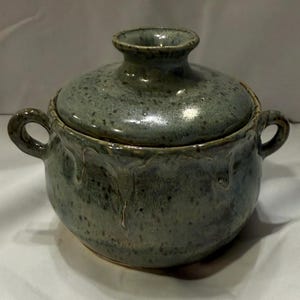 May include: A round ceramic lidded pot with two side handles, featuring a speckled blue-grey glaze with visible drip patterns running down the sides. The lid has a central knob handle, and the overall finish is glossy with a mottled texture.
