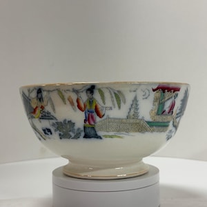 May include: A white porcelain bowl with a pedestal base and a gold rim. The bowl features a colorful Asian-inspired design with figures, pagodas, and foliage. The bowl is likely vintage and suitable for display or serving.