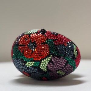 May include: A decorative, oval-shaped object covered in sparkling red, green, purple, and black crystals, arranged in a floral pattern. The object has a small, black, rounded top. The background is a soft, neutral color.