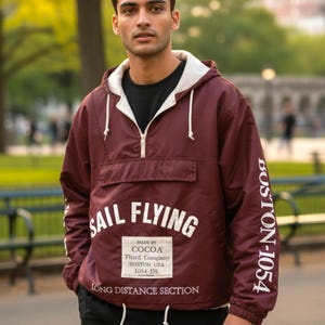 Puede incluir: Chaqueta anorak granate con forro de capucha y cordón blancos. La chaqueta presenta el texto "SAIL FLYING" y "BOSTON-1054" en blanco. Un pequeño parche rectangular dice "MADE BY COCOA Third Company BOSTON USA 1054 DS LONG DISTANCE SECTION".