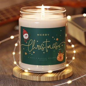 Puede incluir: Una vela de Navidad encendida en un tarro de cristal con una etiqueta verde que presenta una ilustración de un muñeco de nieve y un reno. La etiqueta dice "Merry Christmas" en letras doradas. La vela está sobre un posavasos de madera, rodeada de luces de hadas.