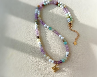 May include: A beaded necklace with a gold-colored initial pendant. The necklace features a variety of colorful beads, including purple, white, green, and turquoise. The necklace is displayed on a neutral-toned surface.